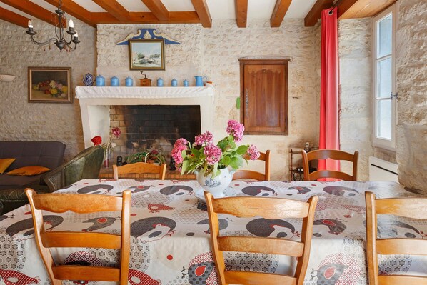 A dining room with a fireplace, wooden chairs, a table with patterned tablecloth, and a vase with flowers.