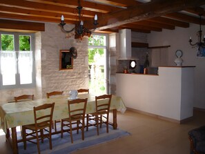 A dining area with a table and chairs, a chandelier, a kitchen area, and a view of greenery outside. - Farm Cottage Between Oysters and Cognac (Antezant-la-Chapelle)
