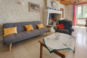 A living room with a sofa, a chair, a table with a tablecloth, a fireplace, and a window with curtains.
