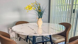 A round marble table with four chairs, a vase with yellow flowers, and a window with vertical blinds.