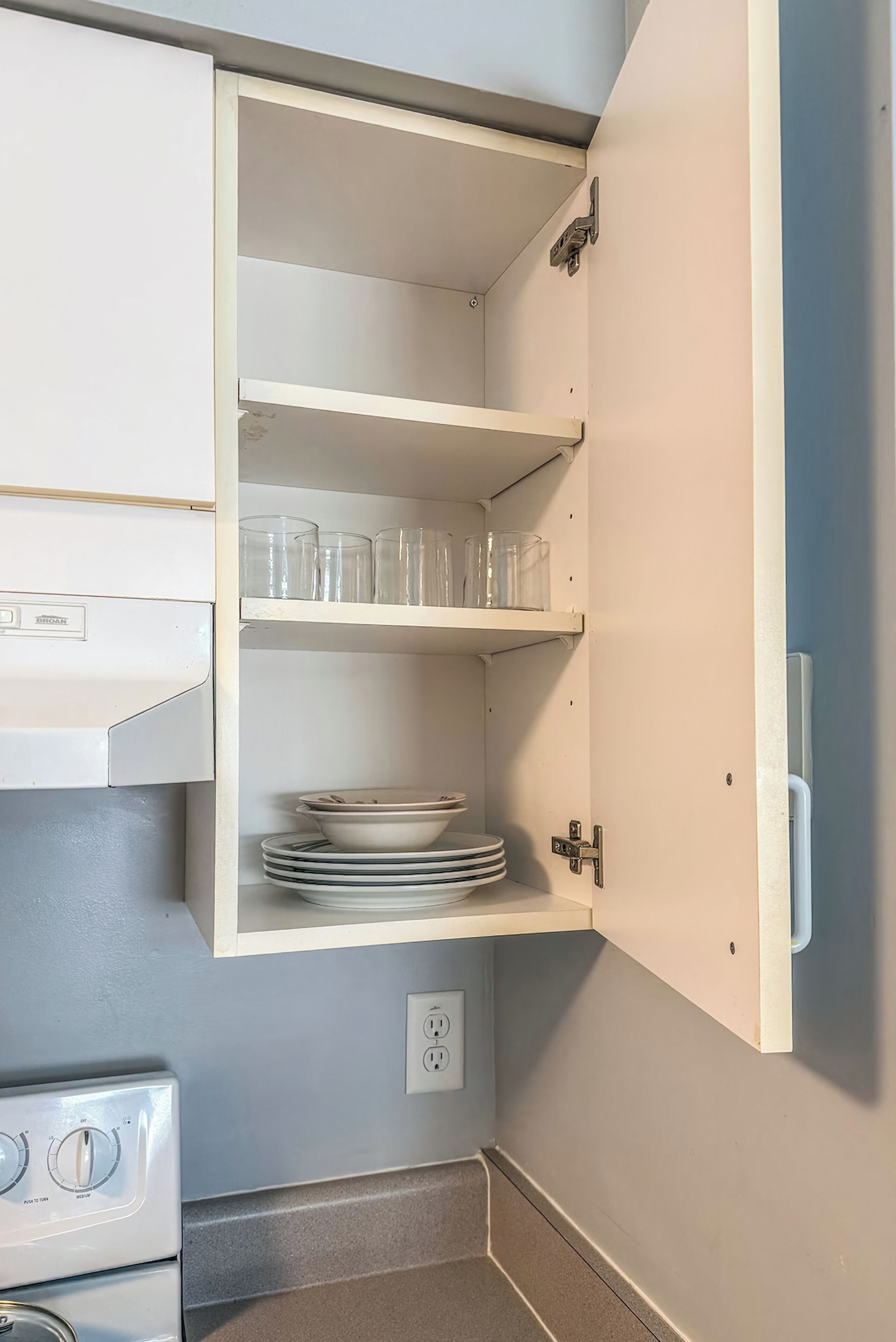 A kitchen with a built-in oven, a stove, and a white cabinet with shelves.
