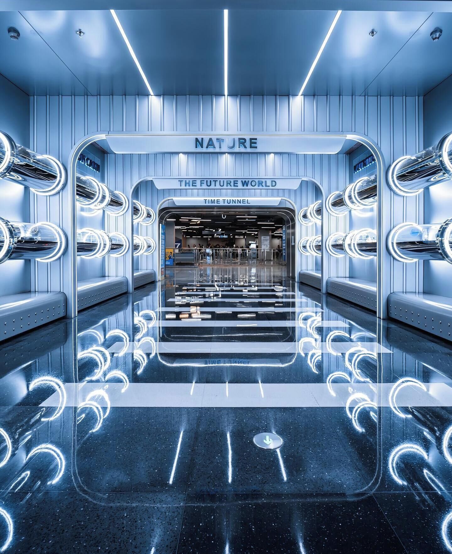 Un interior futurista con una escalera mecánica central que lleva a un nivel inferior, rodeado de superficies reflectantes y luminarias circulares.