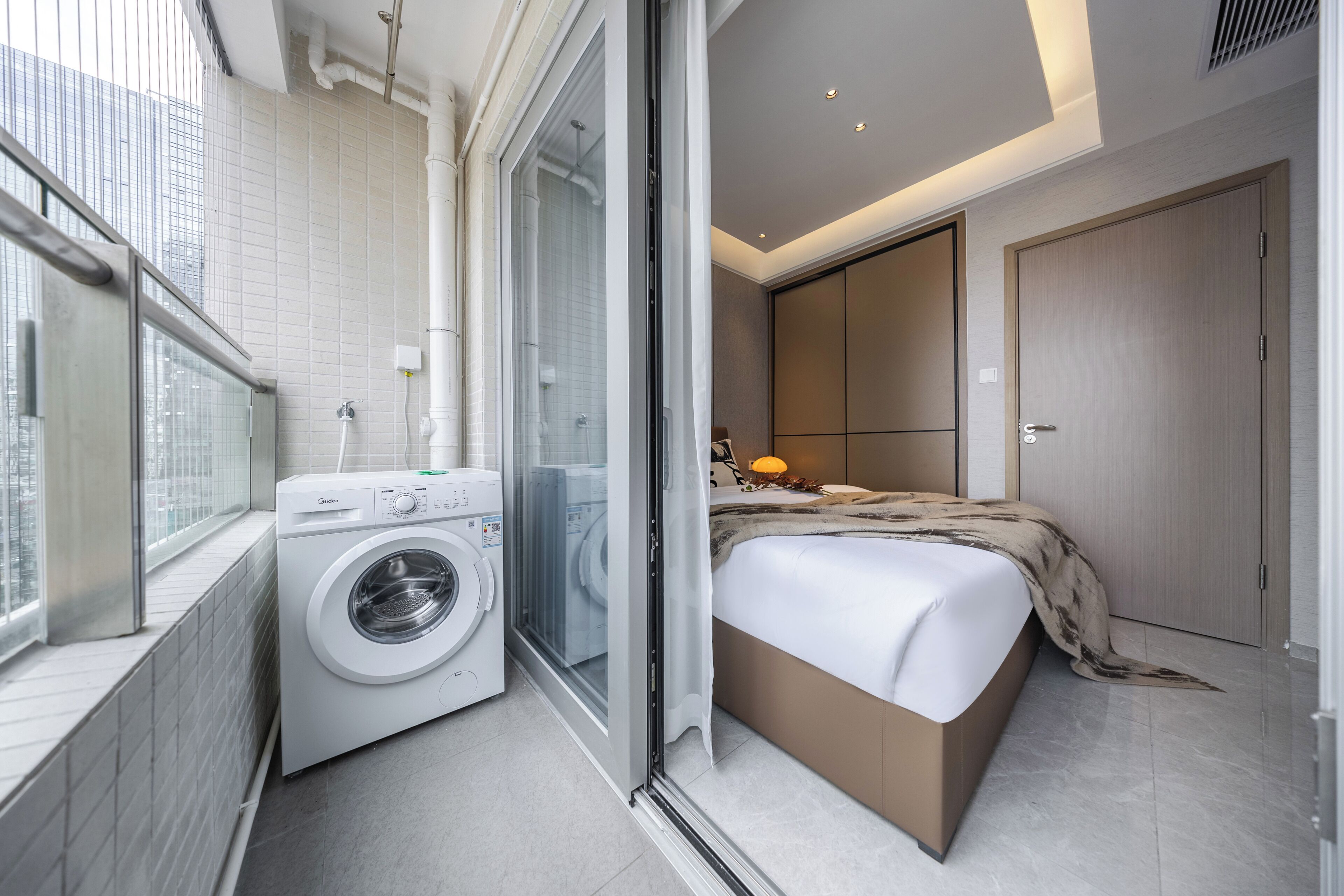 A modern hotel room with a washing machine, a bed with a blanket, a window, and a sliding door.