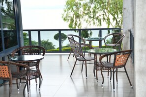 A balcony with wicker furniture and a glass table, overlooking a body of water and greenery. - Golden Rays Hotel (Homa Bay)