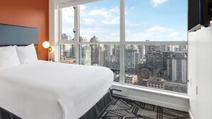 A hotel room with a large bed, a nightstand, a lamp, and a view of the cityscape through large windows.