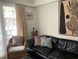 A living room with a black leather sofa, a wicker chair with patterned cushions, a framed picture on the wall, and a large abstract artwork. - Vacation apartment near the beach in a vacation park, sports fields, pet-friendly (De Haan)