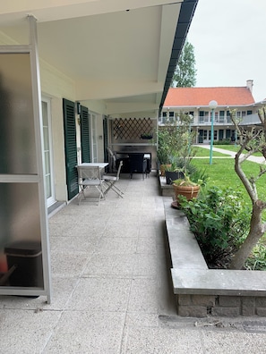 A covered outdoor area with a patio table and chairs, a small garden with potted plants, and a building with a red roof in the background. - Vacation apartment near the beach in a vacation park, sports fields, pet-friendly (De Haan)