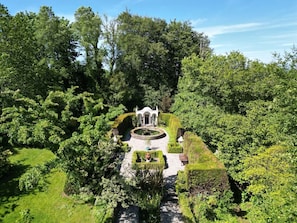 Ein Garten mit einer zentralen Fontäne, umgeben von ordentlich geschnittenen Hecken und Bäumen.