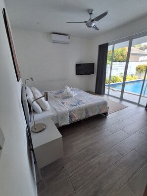 A bedroom with a bed, a desk, a ceiling fan, a TV, and a view of a swimming pool.