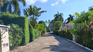 A residential street lined with lush greenery and palm trees, leading to a white car parked on the side. - 2-Bedroom Villa with 2 BTHR, Private Pool, Quiet Beach Just 2-Minute Walk Away (Koh SamuiProvince)