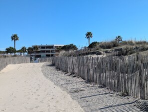 Ein sandiger Strandweg führt zu einem mehrstöckigen Gebäude, umgeben von Palmen und einem Holzzaun.
