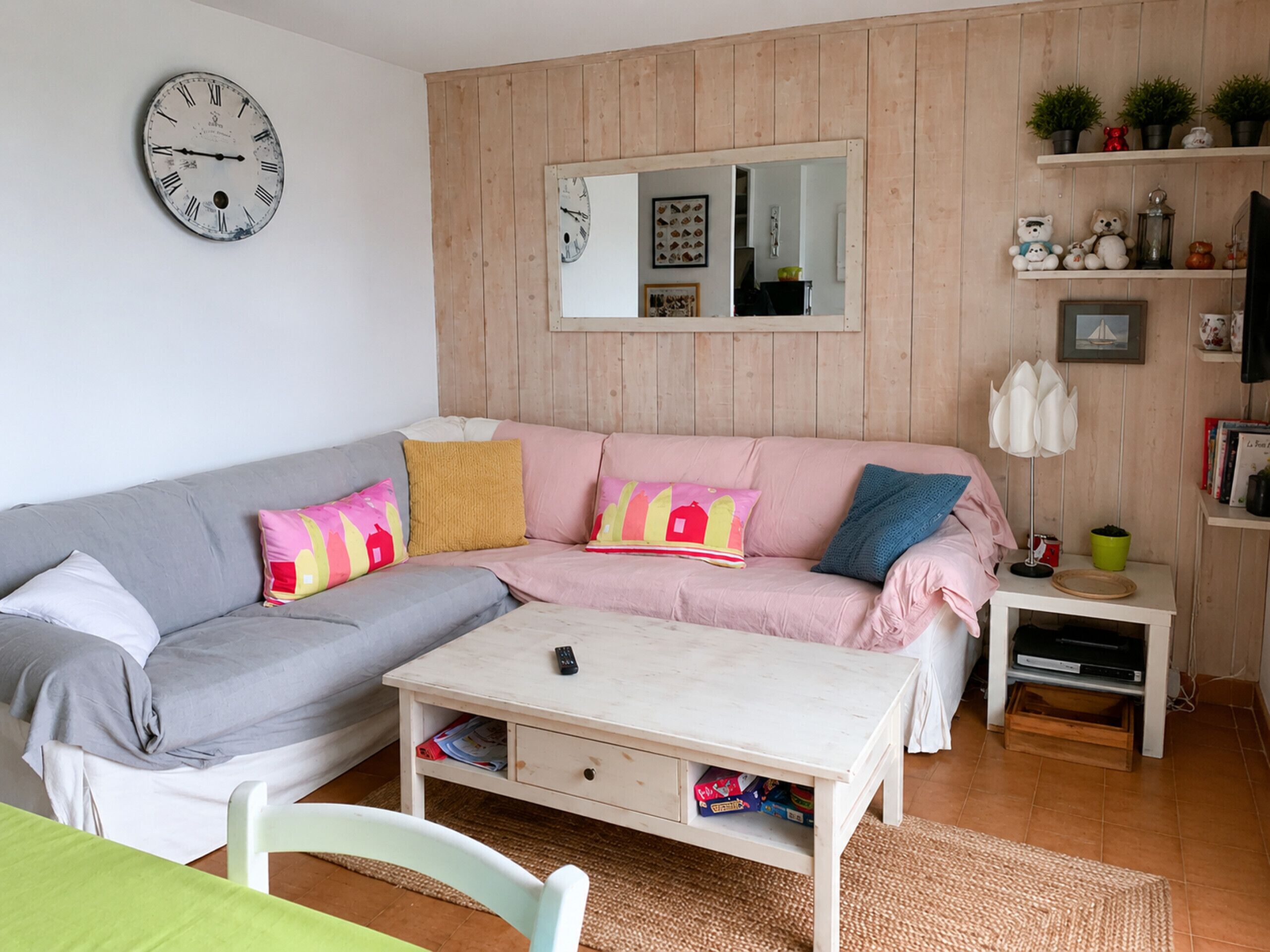 A cozy living room with a sofa, a coffee table, a small table, a clock, a mirror, and a shelf with decorative items.