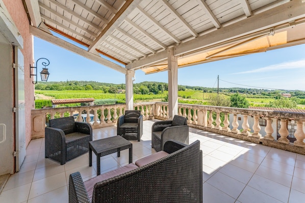A covered outdoor seating area with wicker furniture, a tiled floor, and a view of green fields.