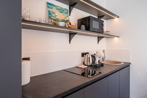 A modern kitchen with black countertops, a microwave, a kettle, and a shelf with kitchenware. - Velvet Quartz Studio | Full Comfort (Rīga)