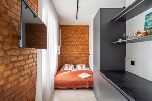 A narrow room with a brick wall, a bed with orange bedding, a black countertop, and a shelf with items. - Velvet Quartz Studio | Full Comfort (Rīga)