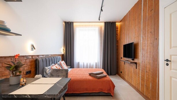Ein gemütliches Zimmer mit Bett, Wandfernseher, Schreibtisch mit Stuhl und einem fenster mit Vorhängen.