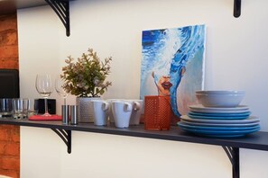 A kitchen counter with a potted plant, white mugs, a red mug, a stack of blue plates, and a painting of a woman. - The Hidden Gem | Smart & Quiet Studio (Riga)