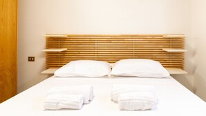 Hotellihuone, jossa on puinen sängynpääty, kaksi tyynyä, valkoisilla lakanoiden peitetty sänky ja kaksi rullalle taitettua pyyhettä sängyllä.