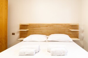 A hotel room with a wooden headboard, two pillows, a bed with white linens, and two rolled towels on the bed. - Golden Sunset - S'Archittu (S'Archittu)