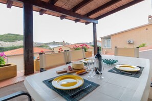 Une table dressée pour deux personnes, avec des assiettes, des verres, une bouteille et un bol, donnant sur une zone côtière avec des bâtiments et de la végétation.
