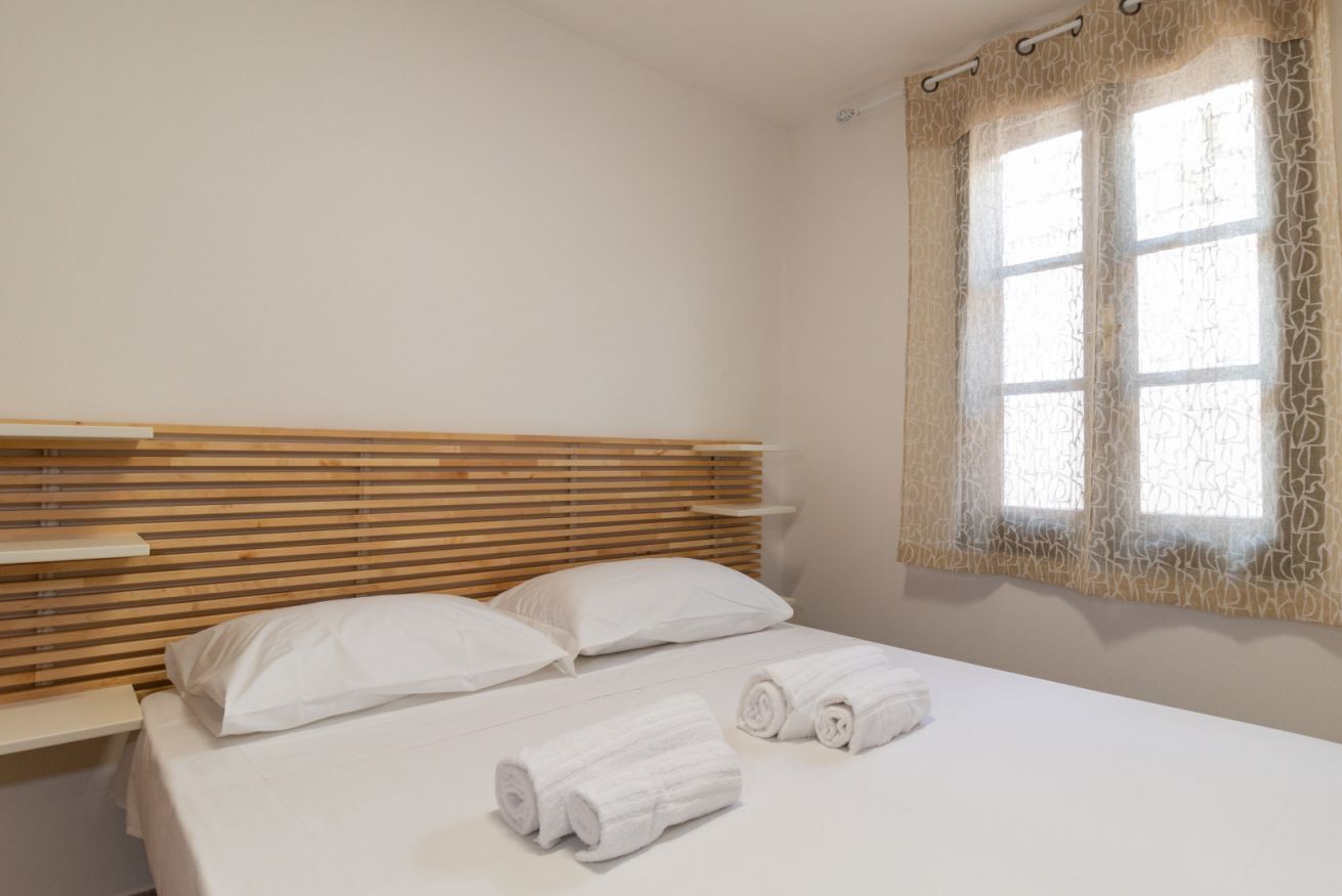 A hotel room with a wooden headboard, a bed with white linens, two rolled towels, and a window with patterned curtains.