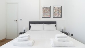 Una cama bien tendida con ropa y toallas blancas, un cabecero oscuro, dos mesitas de noche con lámparas y dos cuadros en la pared.
