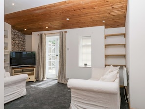 A living room with a television, white furniture, a stone wall, wooden ceiling beams, and a window with blinds. - The Headmasters Quarters (Newton Abbot)