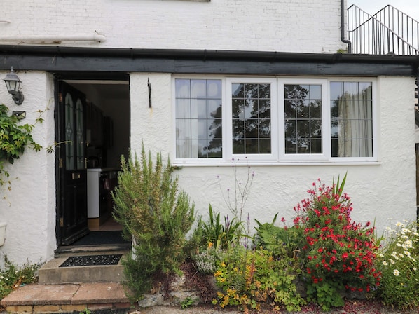 A white building with a black-framed window, a door, and a small garden with various plants and flowers. - The Headmasters Quarters (Newton Abbot)