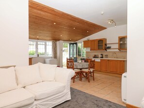 A cozy living room with a white sofa, wooden ceiling, and a view of the kitchen with wooden cabinets and a window.