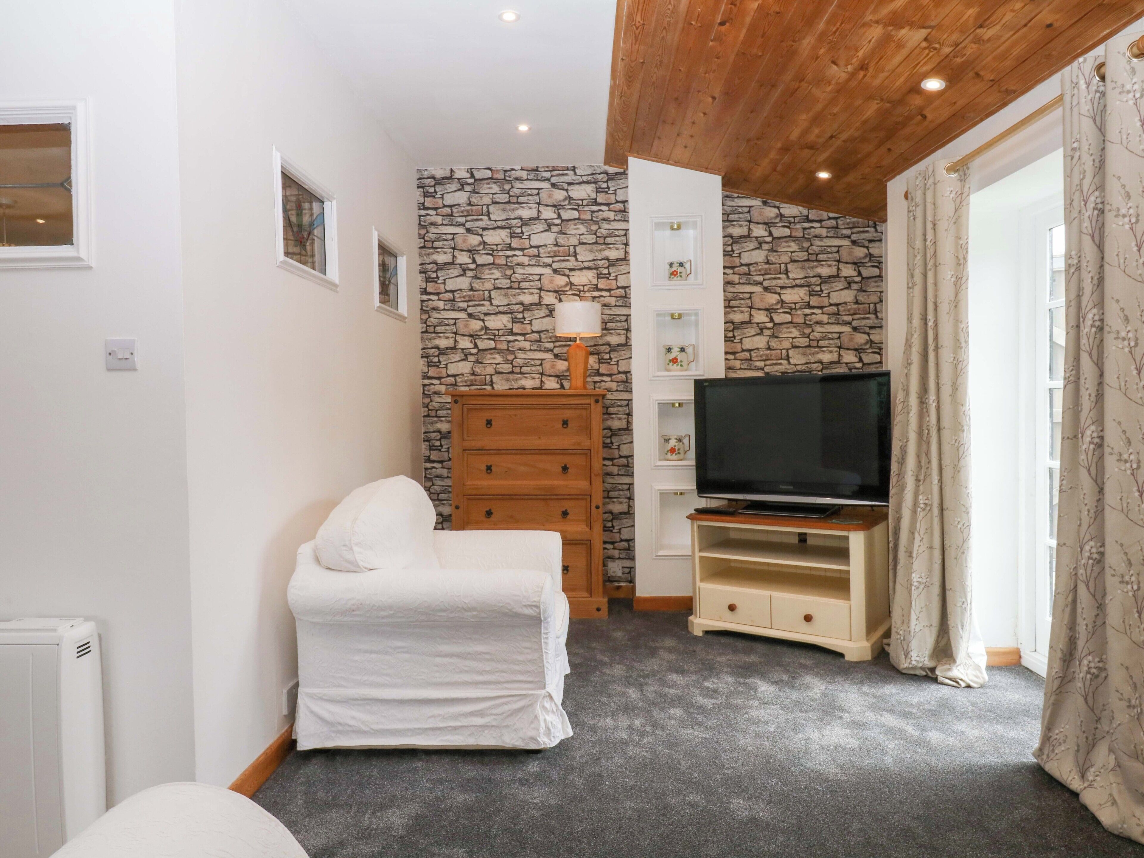 Een kamer met een stenen muur, een witte stoel, een houten dressoir, een televisie op een houten standaard en een radiator.