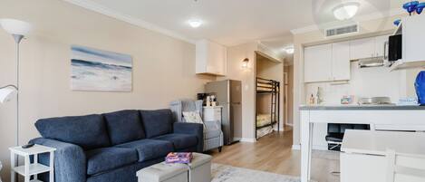 Un salon moderne comprenant un canapé, une table basse, une table d’appoint, un ventilateur de plafond et un coin cuisine avec des placards et un plan de travail.