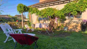 Un jardín con un carretón rojo, sillas de plástico blanco y una mesa lista para comidas al aire libre.