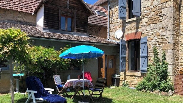 Un patio con una mesa, sillas, un paraguas azul y un edificio de piedra con contraventanas.