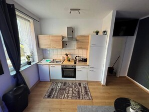 Eine kleine Küche mit Geschirrspüler, Backofen und Spüle. Ein Fenster mit Vorhängen, ein Blumentopf und ein Holzboden.