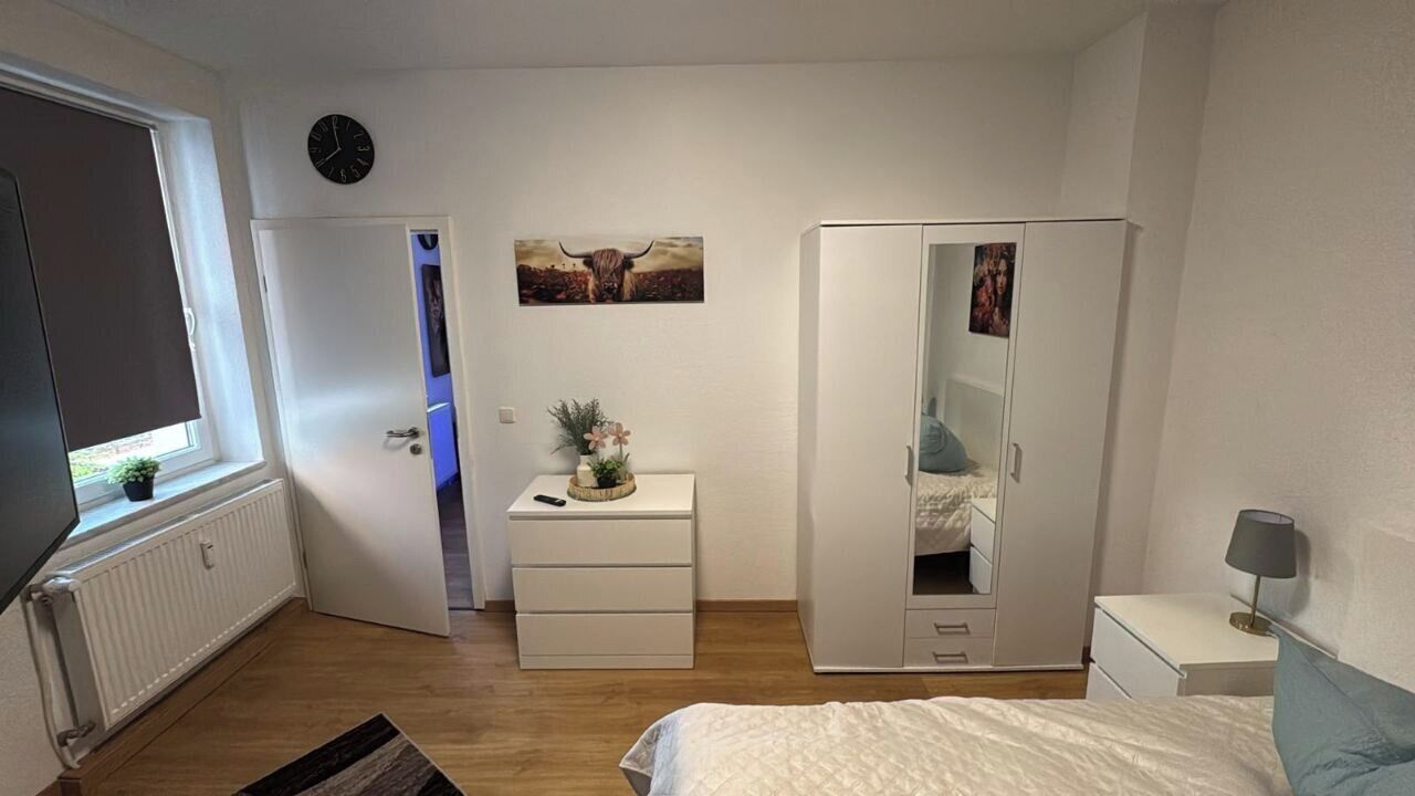 Ein Schlafzimmer mit Bett, Kleiderschrank, Kommode, Spiegel, einer Uhr und einem Fenster.