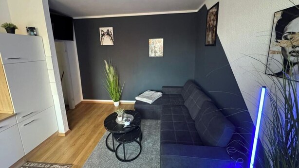 Ein modernes Wohnzimmer mit einem grauen Sofa, einem kleinen schwarzen Couchtisch, einer Zimmerpflanze und Bildern an den Wänden.