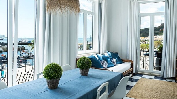 A balcony with a view of the sea, a table with a blue tablecloth, and a sofa with blue cushions. - Floating Over the Sado | Arrábida, Beaches & Town (Setúbal)