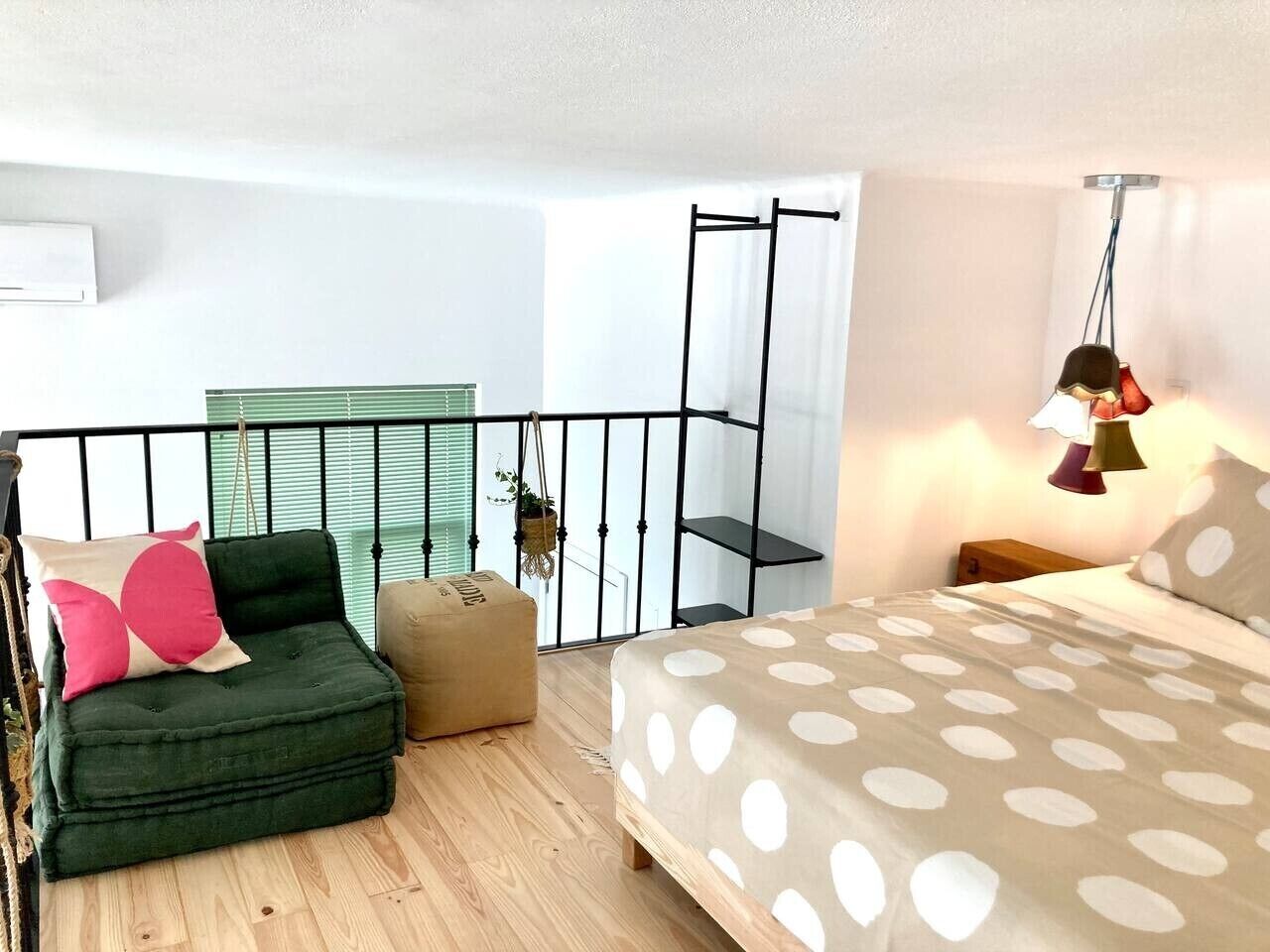 Een slaapkamer met een bed, een groene bank, een klein poufje, een hangplant en een raam met jaloezieën.