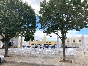 Un espace extérieur aménagé avec des tables et des chaises blanches, des arbres, et un bùtiment en arriÚre-plan.