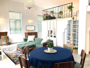 Uma sala de estar com um sofá verde, uma mesa de jantar com toalha azul e uma escada decorada com plantas em vasos.