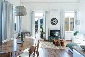 A modern living room with a dining area, a TV, and a large window with curtains. - Waterfront Living | Stunning Sea and Ocean Views (Setúbal)