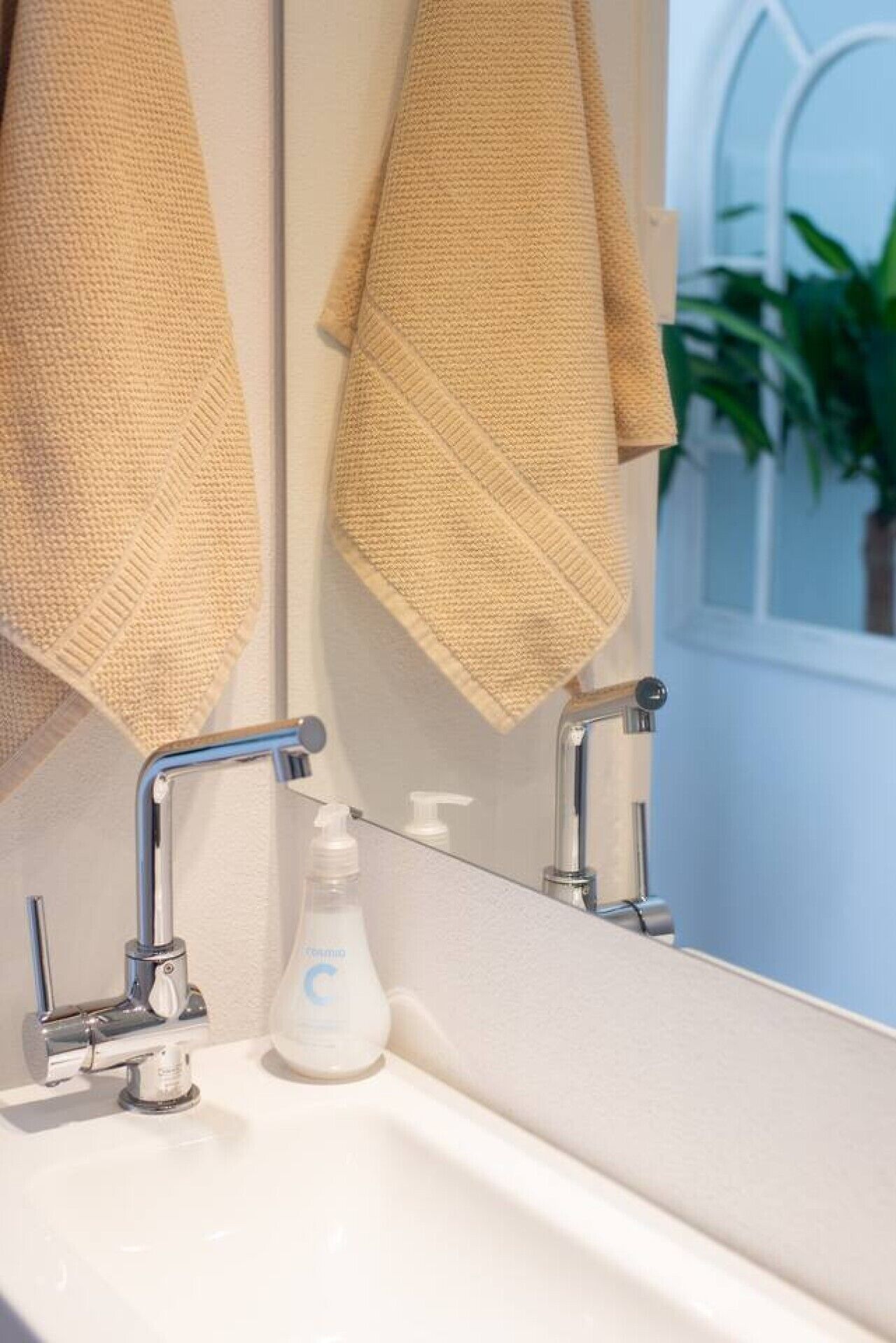 Ein Badezimmer mit einem weißen Waschbecken, zwei chromfarbenen Wasserhähnen, einem Seifenspender und zwei beigen Handtüchern, die an einem Spiegel hängen.