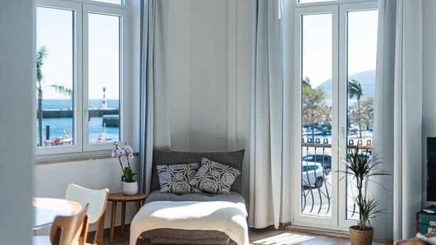 Ein Schlafzimmer mit einem Bett, einem kleinen Tisch, einem Stuhl, einer Topfpflanze und großen Fenstern mit Blick auf das Meer und die Berge.