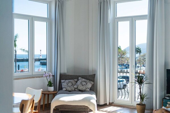A bedroom with a bed, a small table, a chair, a potted plant, and large windows with a view of the sea and mountains. - Waterfront Living | Stunning Sea and Ocean Views (Setúbal)