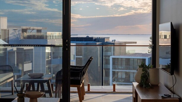 A modern interior with a large window, a dining area with a table and chairs, and a view of the cityscape and water. - Oceanique Penthouse in Leighton Beach (North Fremantle)