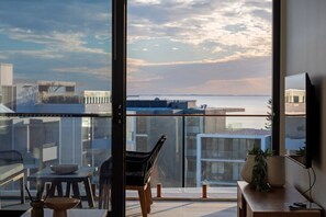 Un intérieur moderne avec une grande fenêtre, un coin repas comprenant une table et des chaises, et une vue sur le paysage urbain et l’étendue d’eau.