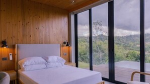 A bedroom with a wooden ceiling, a bed, a nightstand, a chair, and a large window with a view of the outdoors.