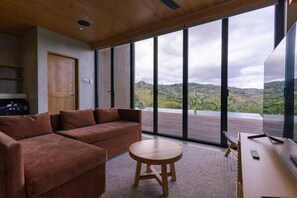 Una habitación moderna con sofá, una pequeña mesa redonda y un ventanal con vistas a las colinas.