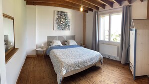 Ein Schlafzimmer mit Holzboden, einem Bett mit gemustertem Überwurf, einem Kopfteil mit geometrischem Muster, einem an der Wand hängenden abstrakten Kunstwerk, einem Fenster mit Vorhängen und einem Heizkörper.