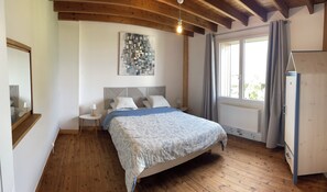 Ein Schlafzimmer mit Holzboden, einem Bett mit gemustertem Überwurf, einem Kopfteil mit geometrischem Muster, einem an der Wand hängenden abstrakten Kunstwerk, einem Fenster mit Vorhängen und einem Heizkörper.